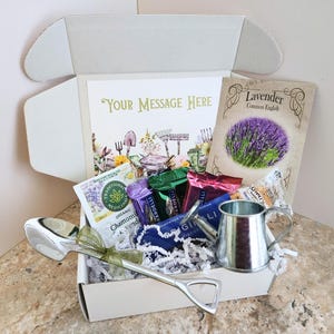 May include: A gift box with a gardening theme, containing a card with the text "Your Message Here", a packet of lavender seeds, tea packets, a small watering can, and a large silver spoon.
