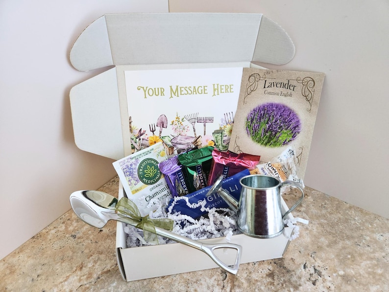 May include: A gift box with a gardening theme, containing a card with the text "Your Message Here", a packet of lavender seeds, tea packets, a small watering can, and a large silver spoon.