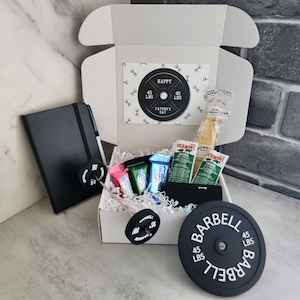 May include: A white gift box with a Father's Day theme, containing a black notebook, snacks, and a barbell weight plate. The plate reads "BARBELL 45 LBS". A card inside the box says "HAPPY FATHER'S DAY".