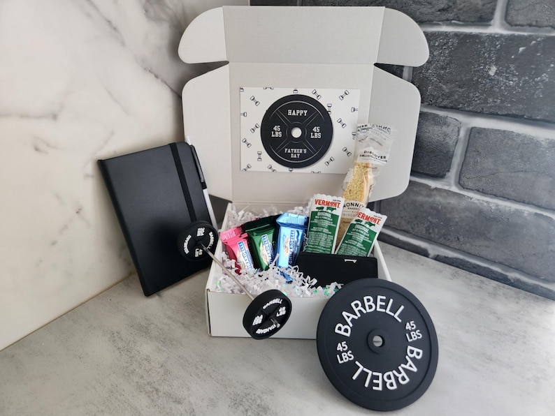 May include: A white gift box with a Father's Day theme, containing a black notebook, snacks, and a barbell weight plate. The plate reads "BARBELL 45 LBS". A card inside the box says "HAPPY FATHER'S DAY".