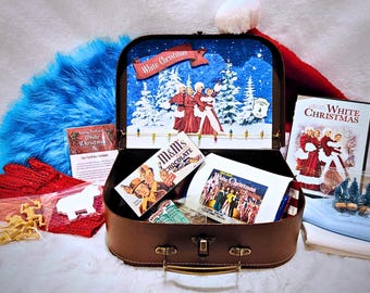 May include: A brown suitcase containing a White Christmas movie DVD, a box of M&M's, a small card, and a printed image. The suitcase interior features a snowy scene with figures in red outfits and the text White Christmas. Accessories include red gloves, blue fabric, and small toy soldiers.