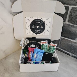 May include: A white gift box with a Father's Day theme, containing a barbell-themed card, snacks, and a small black wallet. The card reads "Happy Father's Day" and features a weight plate design with "45 LBS" on it.