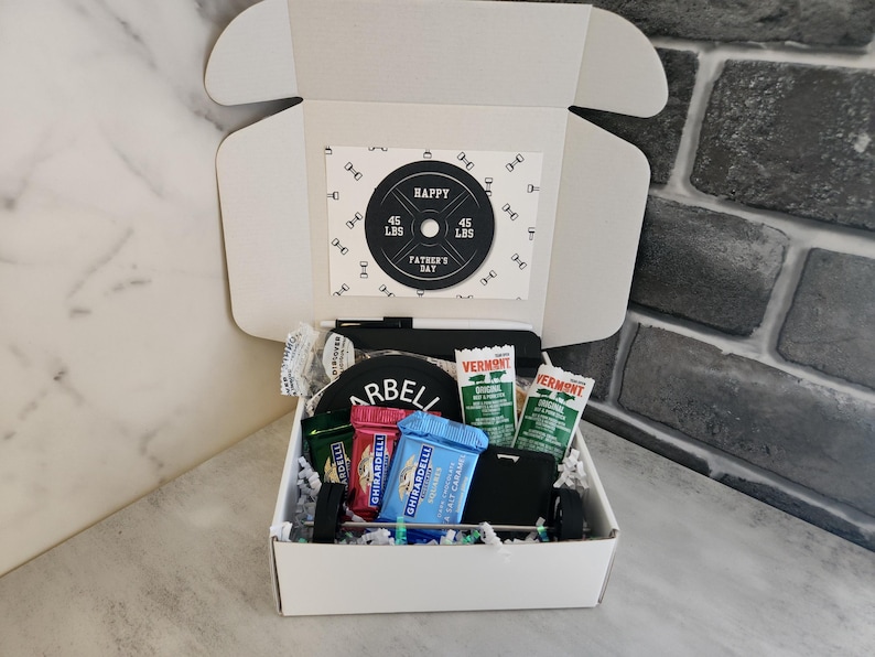 May include: A white gift box with a Father's Day theme, containing a barbell-themed card, snacks, and a small black wallet. The card reads "Happy Father's Day" and features a weight plate design with "45 LBS" on it.