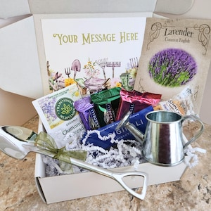 May include: A gift box filled with gardening-themed items. Includes a silver watering can, a large spoon, tea packets, and a packet of lavender seeds. The box also contains a card with the text "Your Message Here".