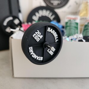 May include: A black barbell weight with white lettering reading "BARBELL", "STANDARD", "20.4 KGS", and "45 LBS". The weight is in front of a white box with other gym-related items.
