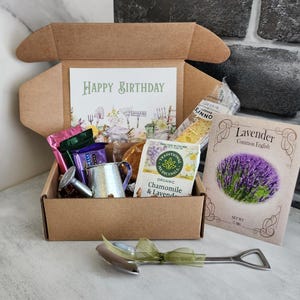May include: A brown gift box filled with various items, including a card that says "Happy Birthday", tea bags, a small metal watering can, and a packet of lavender seeds. A silver shovel-shaped bottle opener with a green ribbon is in front of the box.