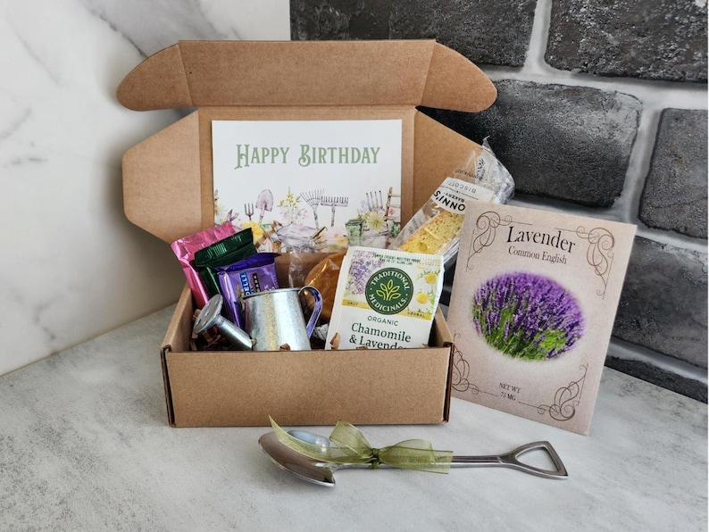 May include: A brown gift box filled with various items, including a card that says "Happy Birthday", tea bags, a small metal watering can, and a packet of lavender seeds. A silver shovel-shaped bottle opener with a green ribbon is in front of the box.