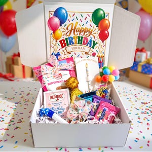 May include: A white cardboard gift box filled with birthday-themed items, including a Red Rose birthday cake tea box, a Ghirardelli chocolate square, a blue party blower, and a card with a candle. The lid features text reading Happy Birthday Jordan.