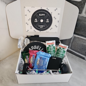May include: A Father's Day gift box with a barbell theme. The box contains a card that says "Happy Father's Day" and a weight plate that says "45 LBS". The box also contains snacks and a wallet.
