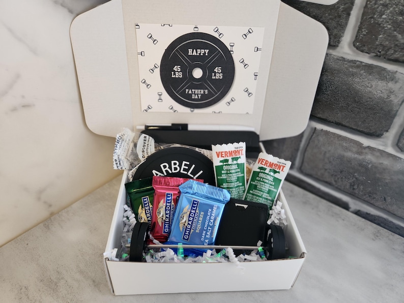 May include: A Father's Day gift box with a barbell theme. The box contains a card that says "Happy Father's Day" and a weight plate that says "45 LBS". The box also contains snacks and a wallet.
