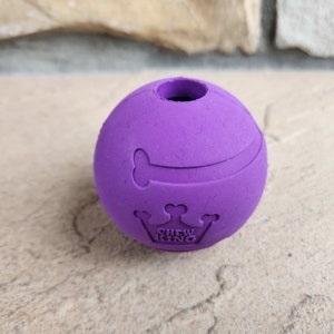 May include: Purple rubber dog chew toy shaped like a ball with a bone and crown design. The toy has the words "Chew King" printed on it.