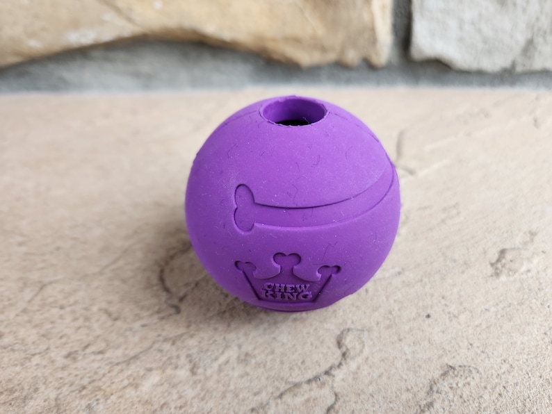 May include: Purple rubber dog chew toy shaped like a ball with a bone and crown design. The toy has the words "Chew King" printed on it.