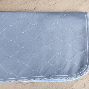 May include: A light blue quilted fabric pad with a white stitched diamond pattern. The pad has a white and blue binding around the edges.