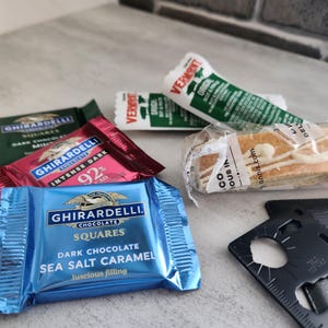 May include: An assortment of packaged snacks and treats, including Ghirardelli chocolate squares in dark chocolate mint, intense dark 92%, and sea salt caramel flavors. Also, a Vermont beef stick and biscotti.
