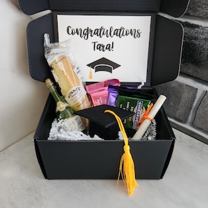 May include: A black gift box with a graduation cap, a bottle of champagne, a baguette, chocolate bars, and a scroll. The box has a card that says "Congratulations Tara!"