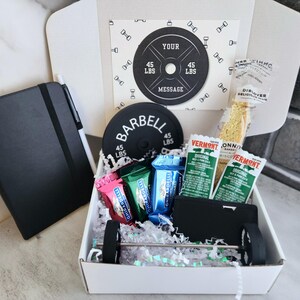 May include: A gift box with a fitness theme, containing a black notebook, pen, barbell-shaped items, and snacks. The box includes a card with the text "YOUR MESSAGE" and a barbell weight of 45 lbs.