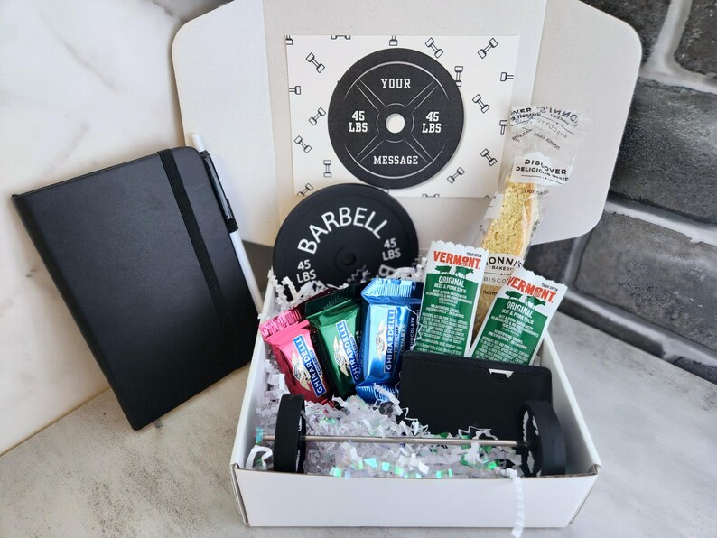 May include: A gift box with a fitness theme, containing a black notebook, pen, barbell-shaped items, and snacks. The box includes a card with the text "YOUR MESSAGE" and a barbell weight of 45 lbs.