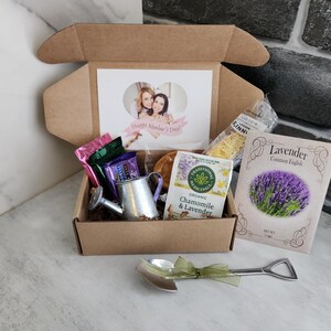 May include: A gift box filled with various items, including a card with a photo and the text "Happy Mother's Day", tea, a miniature watering can, and a packet of lavender seeds. A small shovel-shaped spoon with a green ribbon is also included.