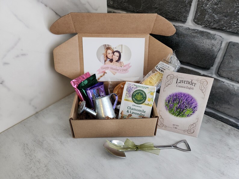 May include: A gift box filled with various items, including a card with a photo and the text "Happy Mother's Day", tea, a miniature watering can, and a packet of lavender seeds. A small shovel-shaped spoon with a green ribbon is also included.