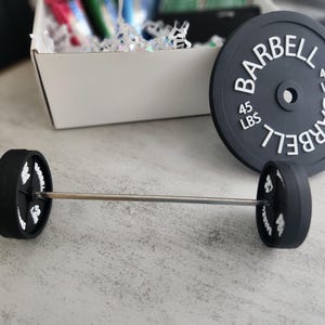 May include: A miniature barbell set with black weights and a silver bar. The weights are labeled "BARBELL" and "45 LBS" in white. A larger weight plate with the same text is also present.