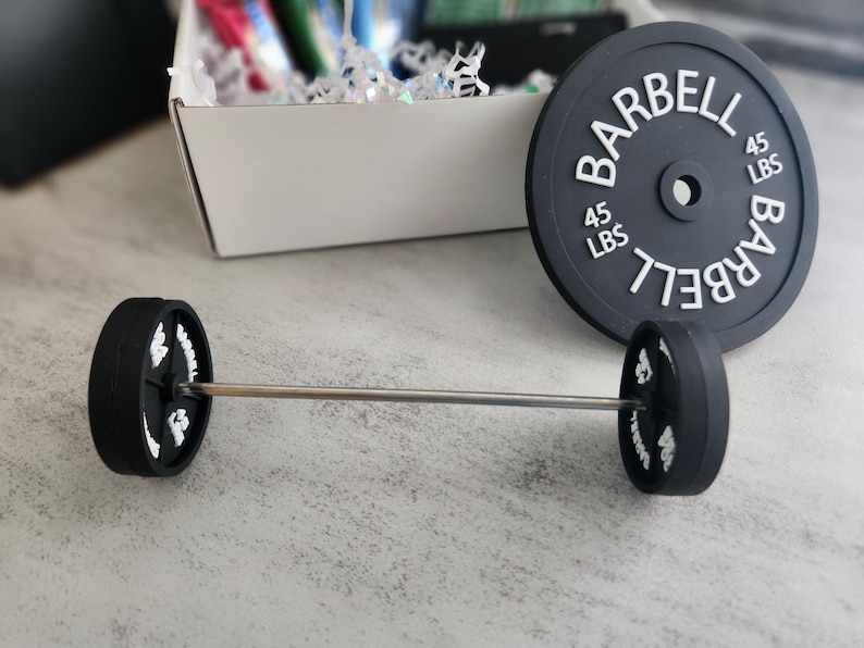 May include: A miniature barbell set with black weights and a silver bar. The weights are labeled "BARBELL" and "45 LBS" in white. A larger weight plate with the same text is also present.