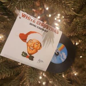 May include: A square Christmas tree ornament featuring the White Christmas album cover by Bing Crosby. The white card shows Bing Crosby wearing a red Santa hat with holly, and a small black vinyl record is attached to the side of the card.