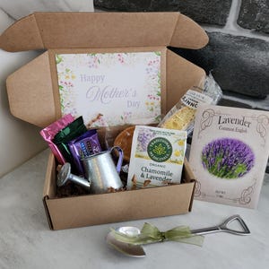 May include: A gift box with a "Happy Mother's Day" card, tea, snacks, and a small watering can. A packet of lavender seeds and a decorative spoon with a green ribbon are also included. The box is brown and the contents are arranged inside.