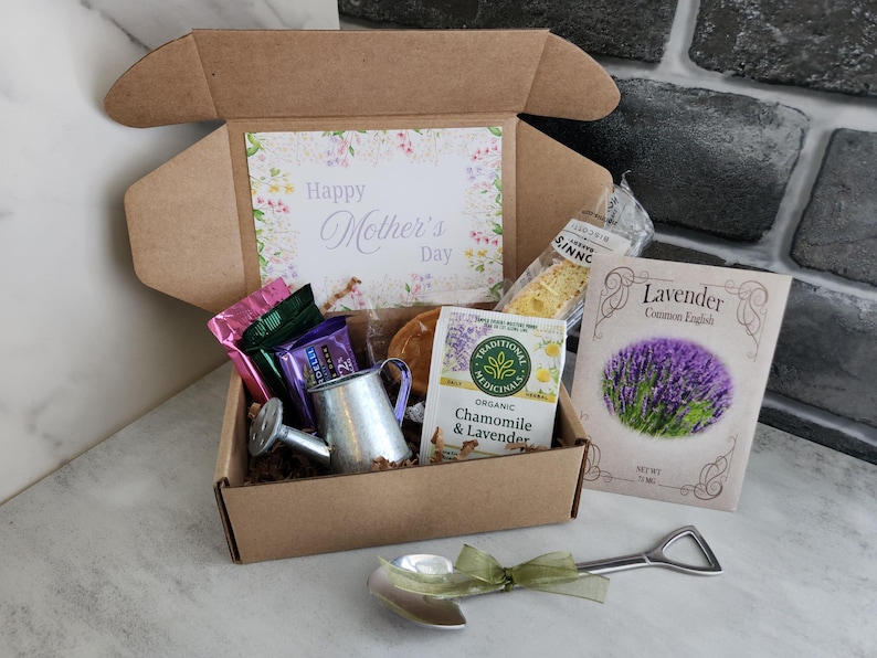 May include: A gift box with a "Happy Mother's Day" card, tea, snacks, and a small watering can. A packet of lavender seeds and a decorative spoon with a green ribbon are also included. The box is brown and the contents are arranged inside.