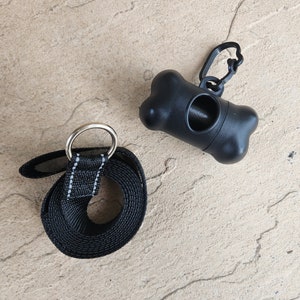 May include: Black dog leash with a silver ring and a black plastic poop bag dispenser with a bone shape and a silver clip.