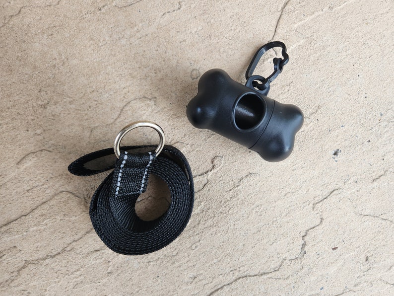 May include: Black dog leash with a silver ring and a black plastic poop bag dispenser with a bone shape and a silver clip.