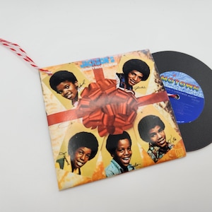 May include: A square ornament featuring the Jackson 5 Christmas Album cover art with a red bow, a red and white string hanger, and a miniature black vinyl record with a blue Motown label attached to the side of the album cover.