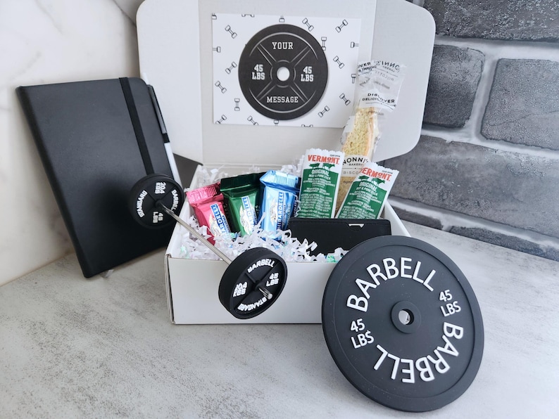 May include: A gift box with a barbell theme, including a black notebook, candy, and a barbell weight plate that reads "BARBELL 45 LBS". The box contains a card with the text "YOUR MESSAGE".