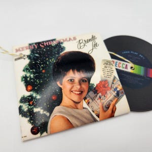 May include: A Brenda Lee Merry Christmas album cover ornament featuring a portrait of the artist with a Christmas tree and a wrapped gift, accompanied by a miniature black vinyl record with a Decca label and a gold hanging string.