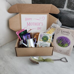 May include: A gift box with a "Happy Mother's Day" card, tea, lavender seeds, and a miniature watering can. The box contains various treats and a decorative spoon with a green ribbon.