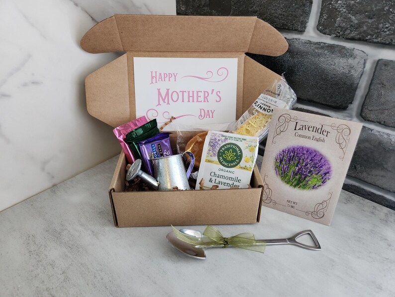 May include: A gift box with a "Happy Mother's Day" card, tea, lavender seeds, and a miniature watering can. The box contains various treats and a decorative spoon with a green ribbon.