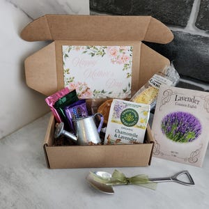 May include: A gift box with a floral "Happy Mother's Day" card, tea, lavender seeds, and a miniature watering can. A shovel-shaped spoon with a green ribbon lies in front of the box. The box is on a marble surface.