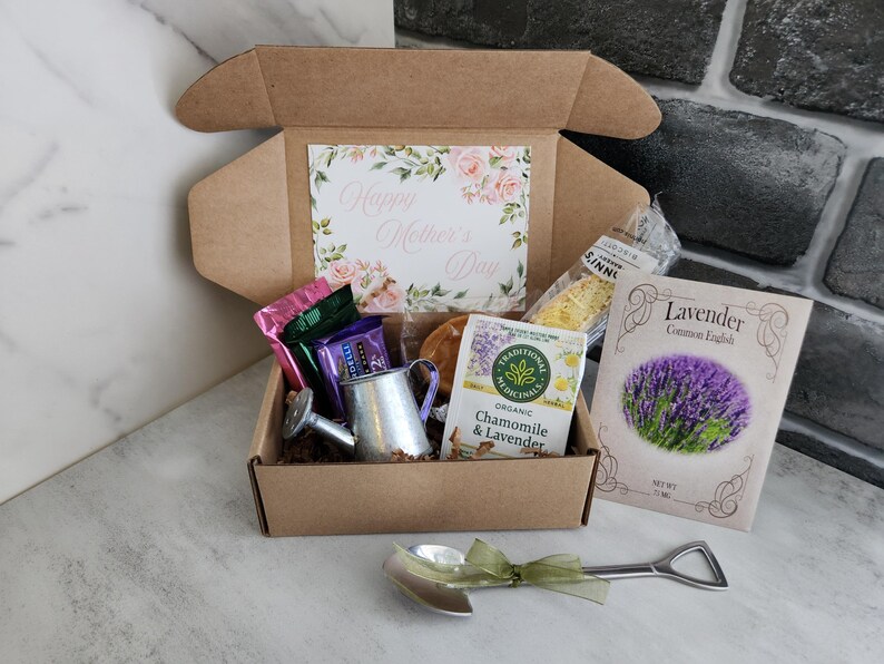 May include: A gift box with a floral "Happy Mother's Day" card, tea, lavender seeds, and a miniature watering can. A shovel-shaped spoon with a green ribbon lies in front of the box. The box is on a marble surface.