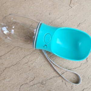 May include: A turquoise blue and clear plastic dog water bottle with a carrying strap. The bottle has a button to dispense water.