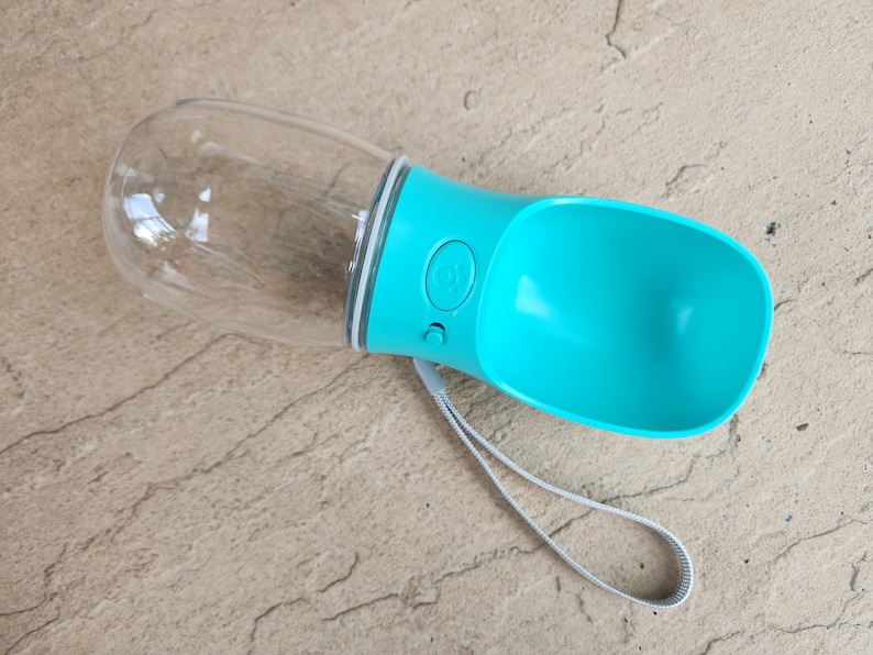 May include: A turquoise blue and clear plastic dog water bottle with a carrying strap. The bottle has a button to dispense water.