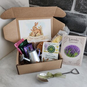 May include: A gift box with a watercolor illustration of two rabbits and the text "Happy Mother's Day." The box contains tea, lavender seeds, a small watering can, and a shovel-shaped spoon with a green ribbon.