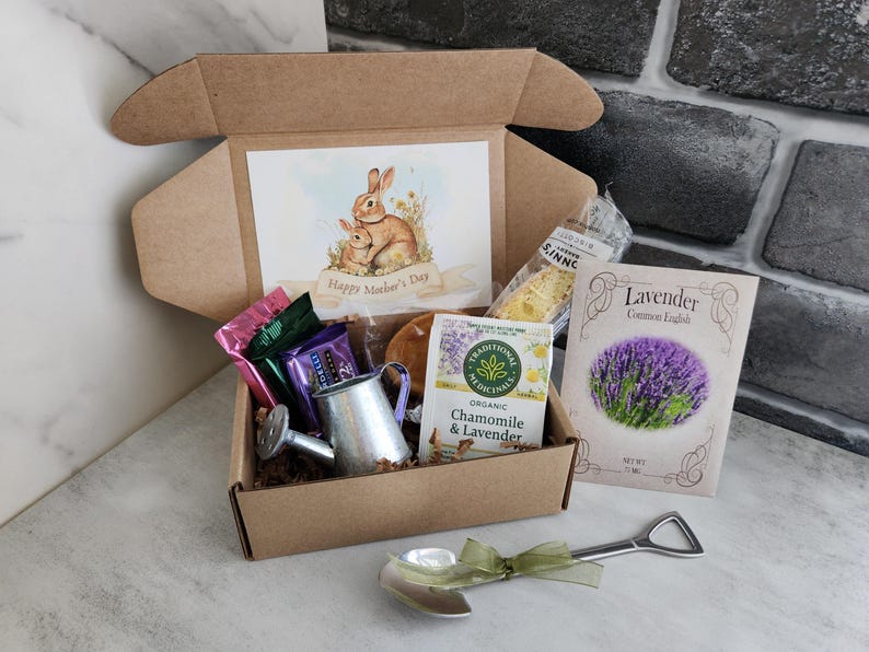 May include: A gift box with a watercolor illustration of two rabbits and the text "Happy Mother's Day." The box contains tea, lavender seeds, a small watering can, and a shovel-shaped spoon with a green ribbon.