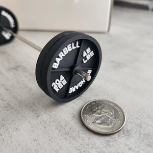 May include: A miniature black barbell with white lettering, including "BARBELL", "45 LBS", and "204 KGS". The barbell is next to a quarter coin, providing a size comparison. The barbell has a silver bar.