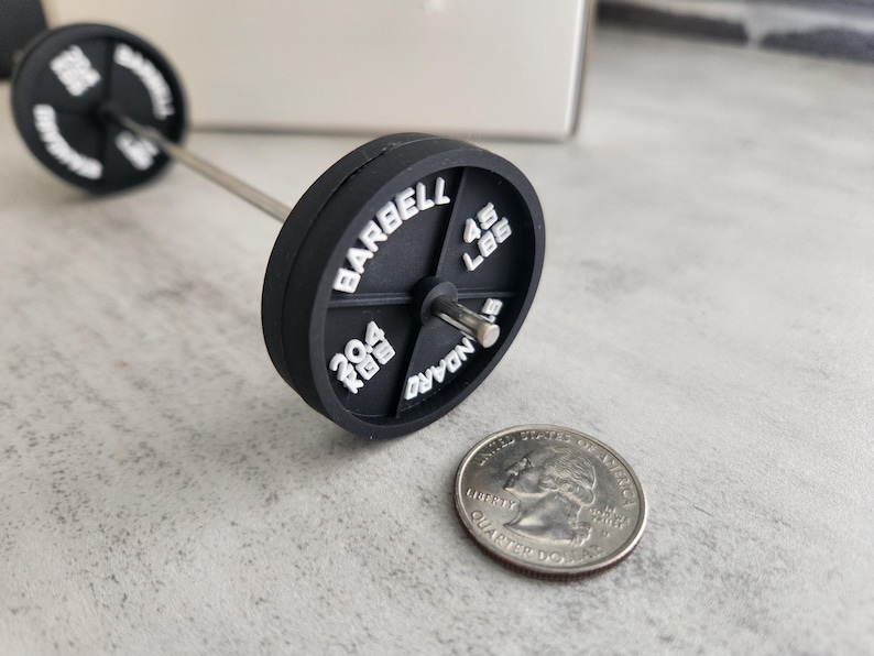 May include: A miniature black barbell with white lettering, including "BARBELL", "45 LBS", and "204 KGS". The barbell is next to a quarter coin, providing a size comparison. The barbell has a silver bar.