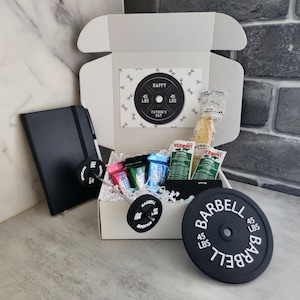 May include: A white gift box with a barbell theme, containing snacks, a black notebook, and a barbell-shaped accessory. The box includes a card that says "HAPPY FATHER'S DAY" and a barbell weight plate that weighs 45 lbs.