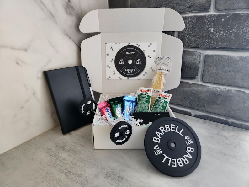 May include: A white gift box with a barbell theme, containing snacks, a black notebook, and a barbell-shaped accessory. The box includes a card that says "HAPPY FATHER'S DAY" and a barbell weight plate that weighs 45 lbs.