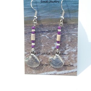 May include: A pair of silver earrings with purple and white beads and a silver seashell charm. The earrings are on a white card with the text "Mi Beach Cape Cod, Cape Cod Wampum & Sand Jewelry, Michele Suvan /shop/ apeCod".