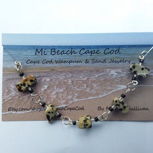 May include: A silver chain bracelet with black beads and brown and white speckled stones. The bracelet is on a card that says "Mi Beach Cape Cod Cape Cod Wampum & Sand Jewelry, Etsy.com/shop/achCapeCod By Mullivan".
