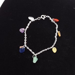 May include: A silver chain bracelet with seven colorful gemstone charms. The charms are various shapes and colors, including purple, red, yellow, green, blue, and orange. The bracelet is displayed on a black surface.