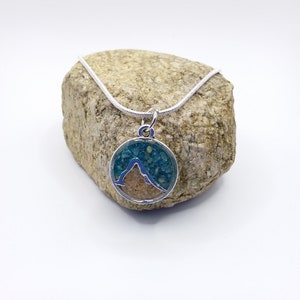May include: A silver chain necklace with a round pendant featuring a mountain silhouette outlined in silver and filled with blue and tan sand.