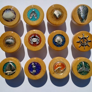 May include: Twelve wooden round ornaments with various nautical themes. Each ornament features a different design, including seashells, a crab, an anchor, a sailboat, and a whale tail. The ornaments are all painted in bright colors and have a glossy finish.